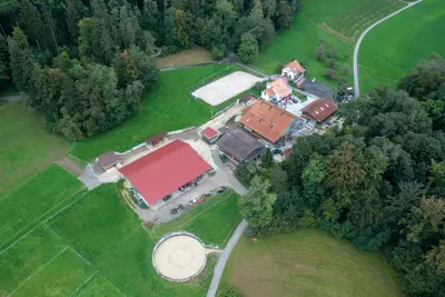 Luftaufnahme des Hofs: Gebäude mit rotem und orangem Dach zwischen Wiesen und Wald, unten ein runder Sandplatz