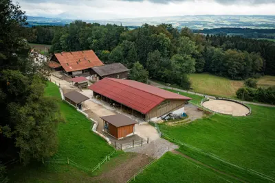 Luftaufnahme: die Hofgebäude mit dem grossen roten Dach, rechts ein runder Sandplatz, dahinter Wald und weite Landschaft