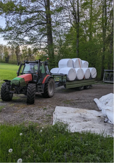 Roter Traktor mit Anhänger und mehreren weiss eingepackten Rundballen unter Bäumen, im Vordergrund liegt weisse Folie