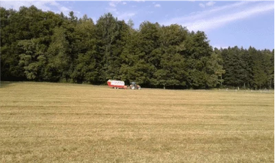 Gemähte Wiese vor einem Waldrand, in der Ferne ein Traktor mit rot-weissem Wagen
