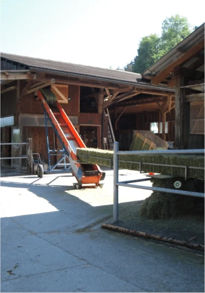 Holzscheune mit einem schrägen Förderband, rechts ein beladener Anhänger