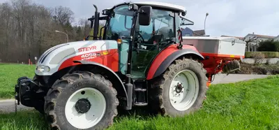 Roter Traktor mit weisser Haube und einem Anbaugerät mit grauem Behälter am Heck, er steht am Rand einer Wiese, dahinter kahle Bäume und Häuser